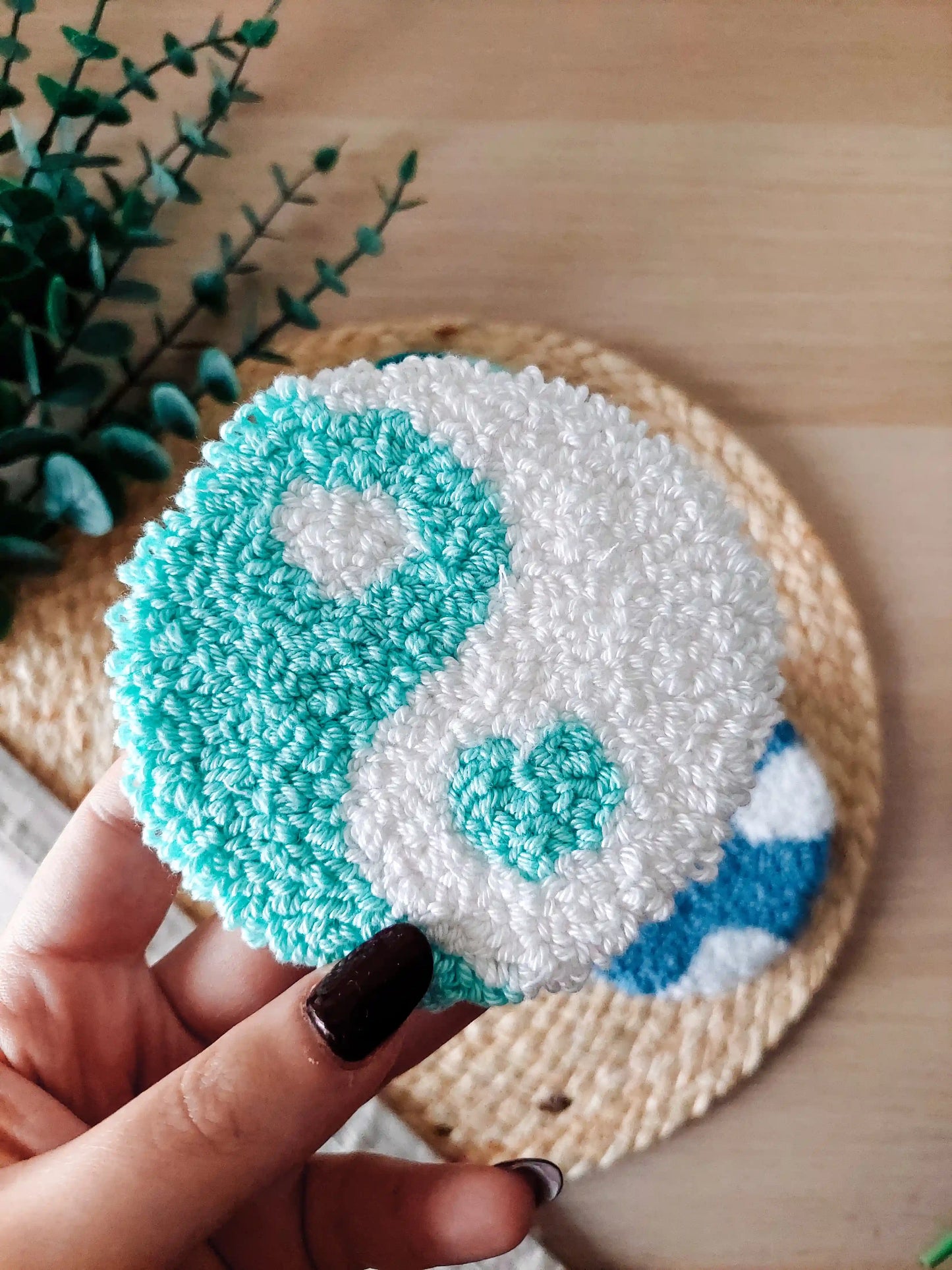 Round mint and white punch needle coaster featuring a yin yang inspired design with a small heart detail, photographed on a woven mat with decorative flowers. 