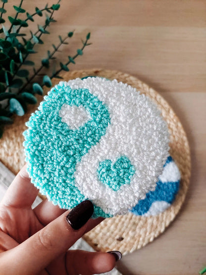 Round mint and white punch needle coaster featuring a yin yang inspired design with a small heart detail, photographed on a woven mat with decorative flowers. 