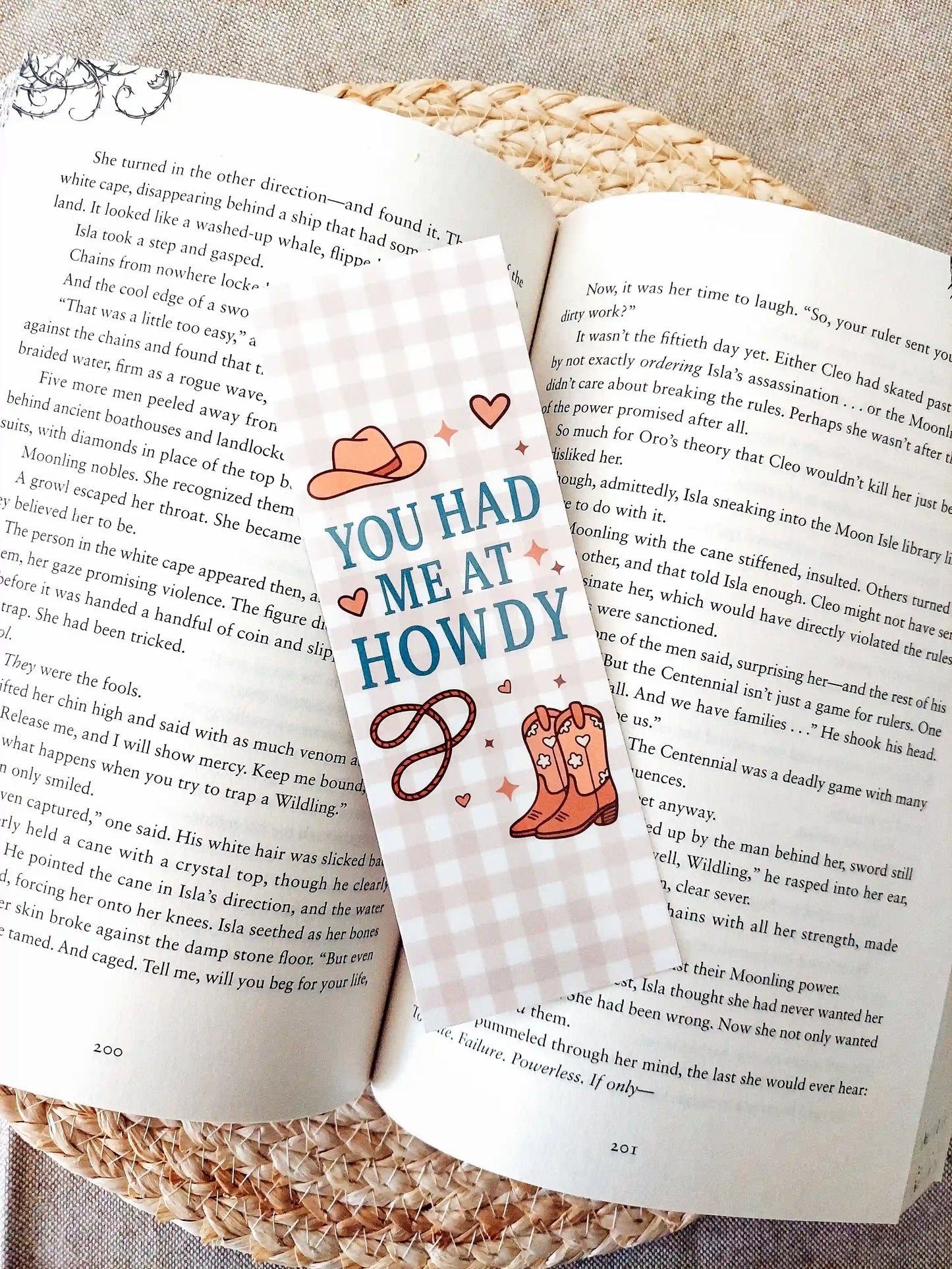 Bookmark with the text "You Had Me at Howdy", featuring a western-themed design with a cowboy hat, boots and rope on a soft gingham background, resting on an open book.