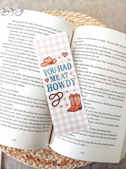Bookmark with the text "You Had Me at Howdy", featuring a western-themed design with a cowboy hat, boots and rope on a soft gingham background, resting on an open book.