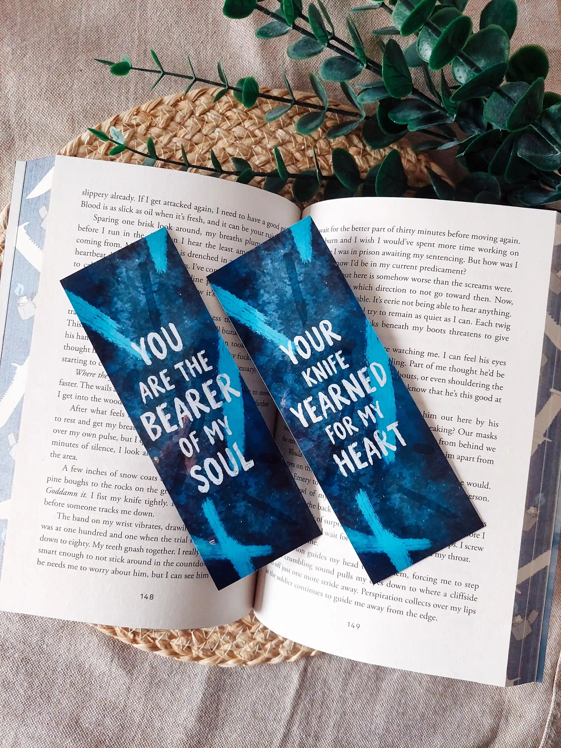 Your Knife, My Heart inspired bookmarks displayed on an open book, featuring a dark blue background with dagger illustrations and white quotes. 