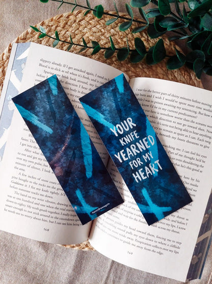Your Knife, My Heart inspired bookmarks displayed on an open book, featuring a dark blue background with dagger illustrations and the quote "Your knife yearned for my heart".